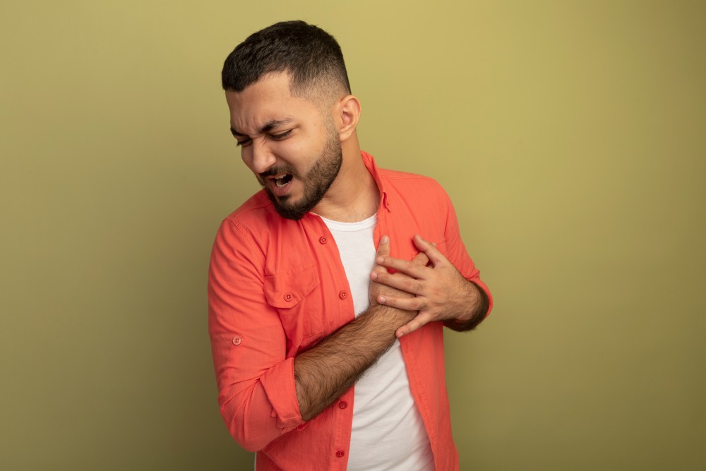young-bearded-man-orange-shirt-holding-hands-his-chest-feeling-pain-standing-light-wall