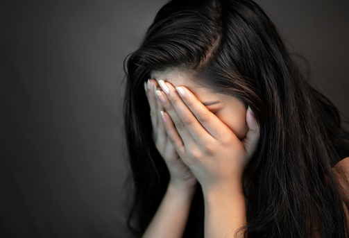 Close-up portrait of alone, stressed young woman sitting in darkness and crying with covering her face with hands.